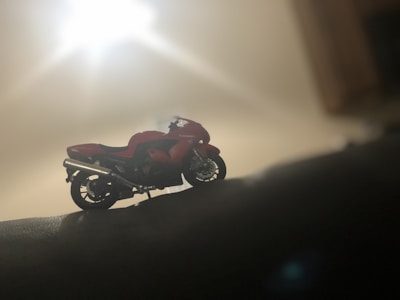 A detailed model of a motorcycle is positioned on a surface in a dimly lit environment. The bike, with its red and black coloration, stands out against the blurred and softly illuminated background. The lighting creates a silhouette effect, enhancing the contours and details of the motorcycle.