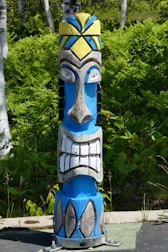 A colorful wooden totem pole with a bold design stands in a lush green forest. The totem features a prominent face with exaggerated features, including large eyes and a wide, toothy grin. It is painted in bright blue, white, yellow, and black colors, adding vibrancy to the natural background of ferns and birch trees.
