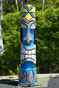 A colorful wooden totem pole with a bold design stands in a lush green forest. The totem features a prominent face with exaggerated features, including large eyes and a wide, toothy grin. It is painted in bright blue, white, yellow, and black colors, adding vibrancy to the natural background of ferns and birch trees.