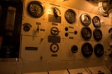 A panel featuring multiple round gauges with letters 'V' and 'A', possibly indicating voltage and amperage. The panel is constructed of metal with various switches, dials, and labels in an industrial setting. Light from an overhead lamp casts a warm glow over the entire setup.