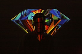 A silhouette of a person stands before a large, colorful, geometric projection resembling a diamond. The image consists of bright, angular patterns in various colors including blue, red, green, and yellow, creating a vibrant and dynamic background against the dark silhouette.