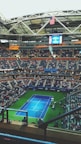 A large tennis stadium filled with spectators features a blue tennis court in the center surrounded by green borders. The stadium's roof is partially visible with an American flag hanging prominently. The seats are mostly filled, indicating a significant event, and a large screen displays information above the players’ entrance.