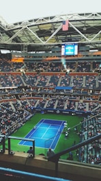 A large tennis stadium filled with spectators features a blue tennis court in the center surrounded by green borders. The stadium's roof is partially visible with an American flag hanging prominently. The seats are mostly filled, indicating a significant event, and a large screen displays information above the players’ entrance.