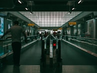 A dimly lit airport walkway with people moving along a travelator. Illuminated signs displaying gate numbers are visible above, while a red X sign indicates the inactive side of the travelator. The space has high ceilings and large windows letting in filtered light.