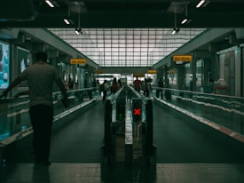 A dimly lit airport walkway with people moving along a travelator. Illuminated signs displaying gate numbers are visible above, while a red X sign indicates the inactive side of the travelator. The space has high ceilings and large windows letting in filtered light.