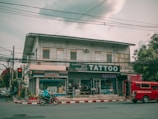 A street view featuring a two-story building with several businesses. The ground floor houses a tattoo parlor with large signage and a shop offering fax and copy services. A red vehicle is parked by the curb while a person rides a motorcycle past on the road. Electrical wires run across the street, and the sky is overcast, casting a muted light over the scene. Trees flank the building on one side.
