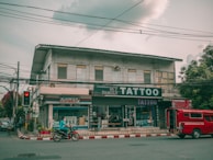 A street view featuring a two-story building with several businesses. The ground floor houses a tattoo parlor with large signage and a shop offering fax and copy services. A red vehicle is parked by the curb while a person rides a motorcycle past on the road. Electrical wires run across the street, and the sky is overcast, casting a muted light over the scene. Trees flank the building on one side.