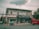 A street view featuring a two-story building with several businesses. The ground floor houses a tattoo parlor with large signage and a shop offering fax and copy services. A red vehicle is parked by the curb while a person rides a motorcycle past on the road. Electrical wires run across the street, and the sky is overcast, casting a muted light over the scene. Trees flank the building on one side.