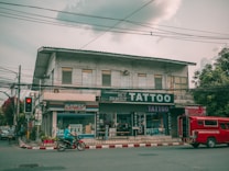 A street view featuring a two-story building with several businesses. The ground floor houses a tattoo parlor with large signage and a shop offering fax and copy services. A red vehicle is parked by the curb while a person rides a motorcycle past on the road. Electrical wires run across the street, and the sky is overcast, casting a muted light over the scene. Trees flank the building on one side.