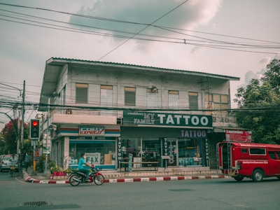 A street view featuring a two-story building with several businesses. The ground floor houses a tattoo parlor with large signage and a shop offering fax and copy services. A red vehicle is parked by the curb while a person rides a motorcycle past on the road. Electrical wires run across the street, and the sky is overcast, casting a muted light over the scene. Trees flank the building on one side.