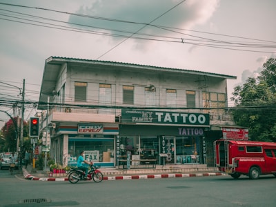 A street view featuring a two-story building with several businesses. The ground floor houses a tattoo parlor with large signage and a shop offering fax and copy services. A red vehicle is parked by the curb while a person rides a motorcycle past on the road. Electrical wires run across the street, and the sky is overcast, casting a muted light over the scene. Trees flank the building on one side.
