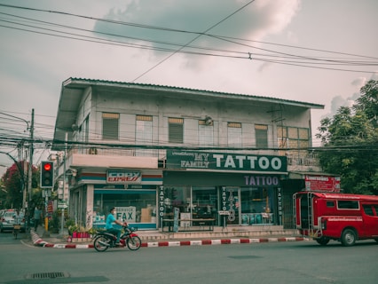 A street view featuring a two-story building with several businesses. The ground floor houses a tattoo parlor with large signage and a shop offering fax and copy services. A red vehicle is parked by the curb while a person rides a motorcycle past on the road. Electrical wires run across the street, and the sky is overcast, casting a muted light over the scene. Trees flank the building on one side.