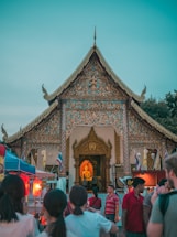 A beautifully ornate temple with intricate carvings and a golden Buddha statue visible through the entrance. The setting is lively with people gathered around, some looking on at the temple while others seem to be engaging in conversation. Colorful banners and tents suggest a festive or market atmosphere. The sky is a soft, serene blue, adding to the cultural ambiance.