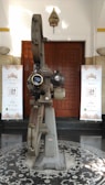 A vintage film projector stands in the center of a patterned floor. It is metallic and intricate, with large reels and lenses. Behind it, wooden double doors are flanked by two banners with a repeating motif, reading "De Majestic - Band Lembang" with a date in 2017. A small decorative hanging is positioned above the doors.