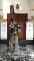 A vintage film projector stands in the center of a patterned floor. It is metallic and intricate, with large reels and lenses. Behind it, wooden double doors are flanked by two banners with a repeating motif, reading "De Majestic - Band Lembang" with a date in 2017. A small decorative hanging is positioned above the doors.