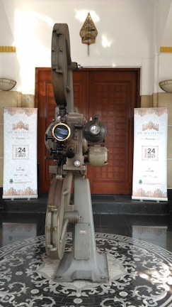 A vintage film projector stands in the center of a patterned floor. It is metallic and intricate, with large reels and lenses. Behind it, wooden double doors are flanked by two banners with a repeating motif, reading "De Majestic - Band Lembang" with a date in 2017. A small decorative hanging is positioned above the doors.