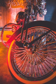 A close-up view of a motorcycle wheel showcasing its intricate, shiny spokes with an orange rim. The background features a warm, ambient glow with a blurred sign and wooden textures, creating a vintage rock-n-roll atmosphere.