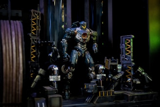A robot-like figure stands in a mechanical setting with intricate details and metallic accents. The figure has a robust design, featuring a central circular element on its chest that glows, arms and legs with visible mechanical joints, and a sleek, armored appearance. Surrounding the figure are various industrial components, including pipes, gears, and structural beams, which contribute to a high-tech environment.