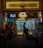 A dimly lit street-side shop with an illuminated sign that reads 'Cheers'. Inside, there are numerous refrigerated display cases stocked with various drinks. Several people are seated outside at tables, having conversations. The atmosphere is casual and social, typical of a small bar or convenience store setting.