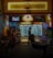 A dimly lit street-side shop with an illuminated sign that reads 'Cheers'. Inside, there are numerous refrigerated display cases stocked with various drinks. Several people are seated outside at tables, having conversations. The atmosphere is casual and social, typical of a small bar or convenience store setting.