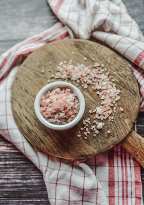 A small white bowl filled with pink Himalayan salt is placed on a round wooden cutting board. The salt is spilled slightly on the board. A red and white checkered cloth is draped around the board, creating a rustic and cozy atmosphere.