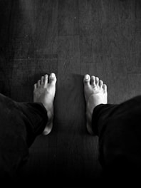 A black and white photograph featuring a person's bare feet on a wooden floor. The feet are positioned apart, with legs in dark pants visible. The floor has a slightly textured surface with visible grains.