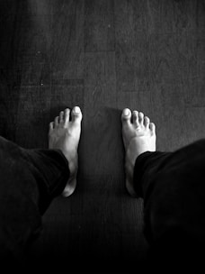 A black and white photograph featuring a person's bare feet on a wooden floor. The feet are positioned apart, with legs in dark pants visible. The floor has a slightly textured surface with visible grains.
