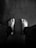 A black and white photograph featuring a person's bare feet on a wooden floor. The feet are positioned apart, with legs in dark pants visible. The floor has a slightly textured surface with visible grains.