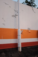 A concrete pole stands in front of a wall painted with horizontal stripes in orange, white, and red. The wall shows signs of wear with several areas where the paint is peeling or damaged. A small part of a plant with green leaves is visible above the top edge of the wall.
