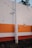 A concrete pole stands in front of a wall painted with horizontal stripes in orange, white, and red. The wall shows signs of wear with several areas where the paint is peeling or damaged. A small part of a plant with green leaves is visible above the top edge of the wall.