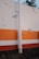 A concrete pole stands in front of a wall painted with horizontal stripes in orange, white, and red. The wall shows signs of wear with several areas where the paint is peeling or damaged. A small part of a plant with green leaves is visible above the top edge of the wall.