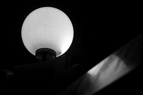 A round, illuminated street lamp is positioned against a dark background, casting a soft glow. Below the lamp, a reflective surface suggests a metallic handrail or structure running diagonally across the frame.