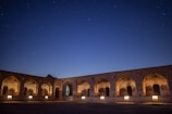 An ancient brick structure with arched doorways and windows is illuminated by warm lights against a backdrop of a starry night sky.