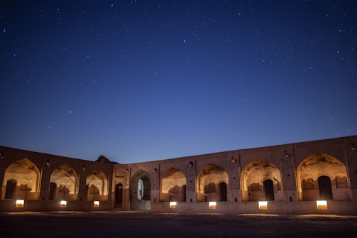 An ancient brick structure with arched doorways and windows is illuminated by warm lights against a backdrop of a starry night sky.