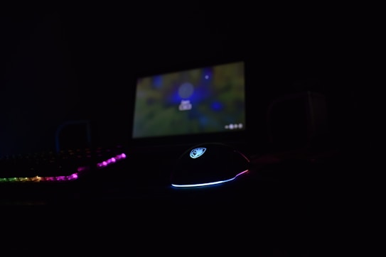 A dimly lit setup featuring a gaming mouse with RGB lighting, a keyboard with backlit keys, and a laptop displaying a blurred background screen. The scene is predominantly dark, with the only sources of light coming from the electronic devices.