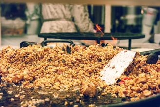 A large serving of cooked rice mixed with various ingredients, including chunks of meat and vegetables, is spread over a wide, shallow cooking pan. A metal spatula rests on top of the rice, suggesting that the food is being prepared or served. In the background, a blurred figure stands near a stove with visible flames.