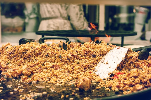 A large serving of cooked rice mixed with various ingredients, including chunks of meat and vegetables, is spread over a wide, shallow cooking pan. A metal spatula rests on top of the rice, suggesting that the food is being prepared or served. In the background, a blurred figure stands near a stove with visible flames.