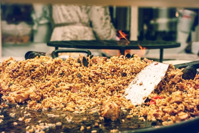 A large serving of cooked rice mixed with various ingredients, including chunks of meat and vegetables, is spread over a wide, shallow cooking pan. A metal spatula rests on top of the rice, suggesting that the food is being prepared or served. In the background, a blurred figure stands near a stove with visible flames.