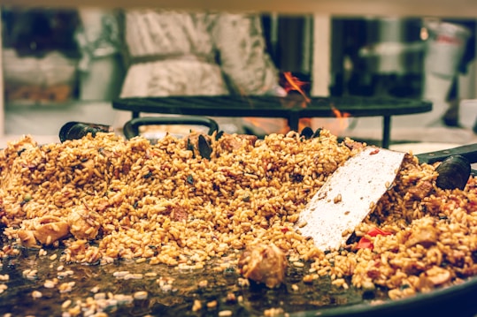 A large serving of cooked rice mixed with various ingredients, including chunks of meat and vegetables, is spread over a wide, shallow cooking pan. A metal spatula rests on top of the rice, suggesting that the food is being prepared or served. In the background, a blurred figure stands near a stove with visible flames.