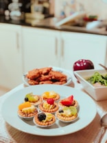 A plate of small fruit tarts topped with various fruits such as kiwi, raspberry, and blackberry, is placed on a table. In the background, there is a bowl of crispy breaded items and a plate containing green vegetables in a broth. The setting appears to be a kitchen with a countertop and kitchen utensils visible.