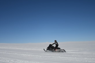 A snowmobile speeding through a snowy landscape under a clear blue sky.