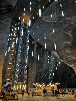 A large, cavernous space resembling a salt mine or underground chamber, featuring tall walls with illuminated vertical light fixtures. The walls have a textured, rock-like surface, appearing dark with swirling patterns. In the foreground, a group of people stands near a modern, circular bar structure with wooden accents and bright overhead lighting. The overall setting combines industrial and natural elements, creating an intriguing underground atmosphere.