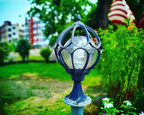 A close-up view of a decorative outdoor lamp post set against a blurred background of greenery and buildings. The lamp has a spherical design with a spiral light bulb enclosed in a decorative metal cage. Surrounding the lamp are green plants and flowers, with hints of red and white stripes visible in the background.