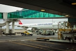 A Turkish Airlines plane is parked at an airport gate with a glass boarding bridge connected to it. Various airport service vehicles, including a yellow and black taxi, are positioned nearby under a cloudy sky.