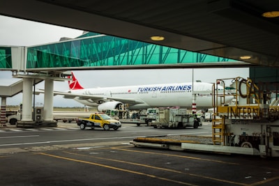 A Turkish Airlines plane is parked at an airport gate with a glass boarding bridge connected to it. Various airport service vehicles, including a yellow and black taxi, are positioned nearby under a cloudy sky.