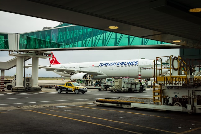A Turkish Airlines plane is parked at an airport gate with a glass boarding bridge connected to it. Various airport service vehicles, including a yellow and black taxi, are positioned nearby under a cloudy sky.