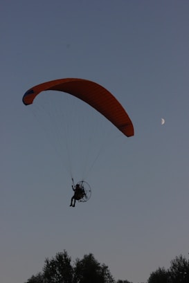 A person is flying in a powered paraglider with a large orange canopy against a dusky sky. The moon is visible in the background, providing a serene atmosphere. Below, the silhouette of treetops can be seen.