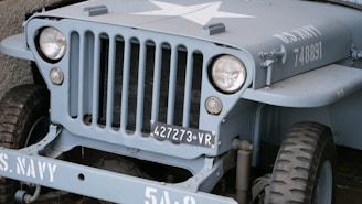 A vintage military jeep painted in grey-blue featuring a prominent white star on the hood. The vehicle has a distinctive grille with vertical slots, round headlights, and a visible license plate marked with '427273 VR'. It is marked with 'U.S. NAVY 748891' on the side.