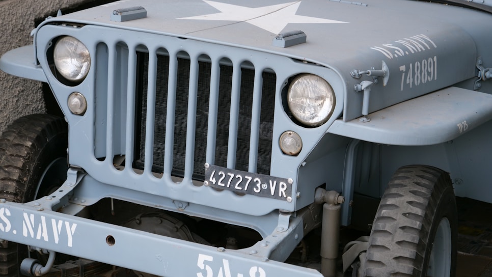 A vintage military jeep painted in grey-blue featuring a prominent white star on the hood. The vehicle has a distinctive grille with vertical slots, round headlights, and a visible license plate marked with '427273 VR'. It is marked with 'U.S. NAVY 748891' on the side.