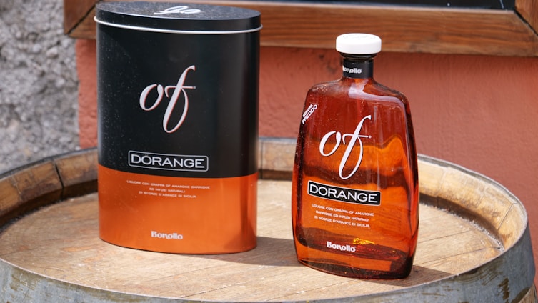 A dark cylindrical tin and a bottle of liqueur rest on a wooden barrel, both labeled 'of DORANGE' by Bonollo. The bottle is amber-colored, and the surroundings include a rough-textured wall in the background.